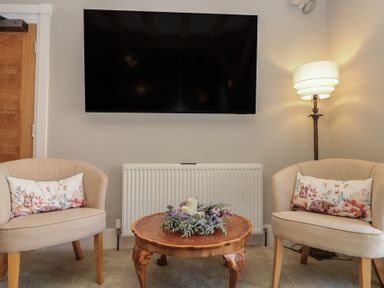 A sitting area with two beige chairs with animal print cushions a wooden coffee table with candles and flowers a floor lamp and a wall mounted television at The Barn Helperby near Boroughbridge