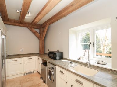 A kitchen with wooden beams on the ceiling white cabinets a countertop microwave washing machine sink and two pet bowls by a window at The Barn Helperby near Boroughbridge