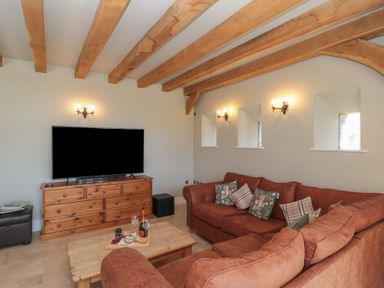 A living room with wooden beams on the ceiling a large flat screen tv on a wooden dresser and a brown sofa with patterned cushions at The Barn in Helperby near Boroughbridge