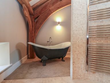 A bathroom with a clawfoot bathtub under wooden beams and a tiled wall with a heated towel rail at The Barn Helperby near Boroughbridge