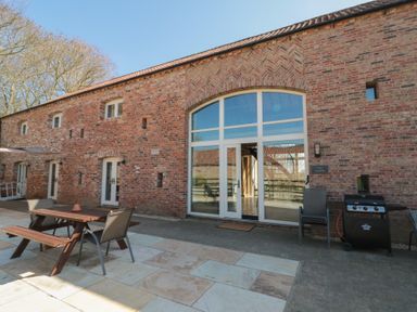 An outdoor patio area with wooden picnic table and chairs next to a brick building with large arched window and doors at The Barn in Helperby near Boroughbridge