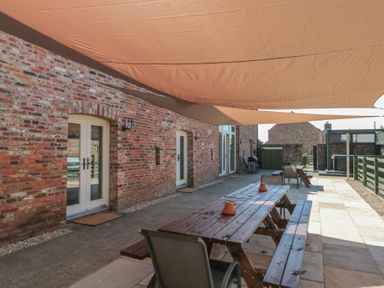 An outdoor patio area with a wooden picnic table and chairs against a brick building with white doors at The Barn in Helperby near Boroughbridge