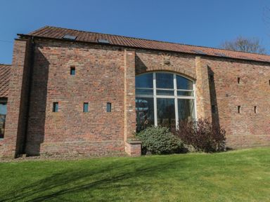 A brick building with a large arched window and small rectangular windows surrounded by grass at The Barn Helperby near Boroughbridge