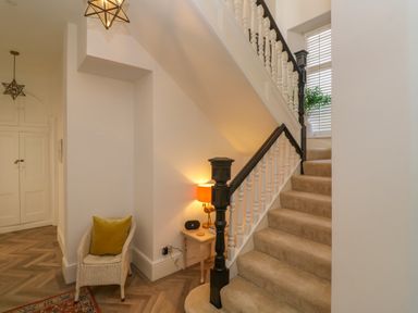 A staircase with beige carpet and white railing a chair with a green cushion a small table with an orange lamp and a decorative light fixture in the hallway at Hindon Hall in Lelant near Carbis Bay