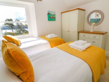 A bedroom with two single beds with yellow blankets and pillows a white wardrobe and a white dresser with a round mirror and a window showing trees and a water view at Hindon Hall in Lelant near Carbis Bay