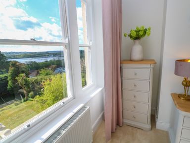 A corner of a bedroom with a window showing a view of trees and water a small chest of drawers with a vase of flowers and a bedside table with a lamp at Hindon Hall in Lelant near Carbis Bay