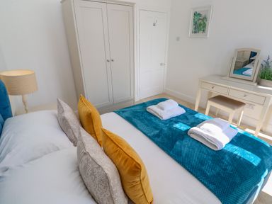 A bedroom with a bed with multiple pillows and folded towels on a blue blanket a wardrobe a dressing table with a mirror and potted plant and a lamp on a bedside table at Hindon Hall in Lelant near Carbis Bay