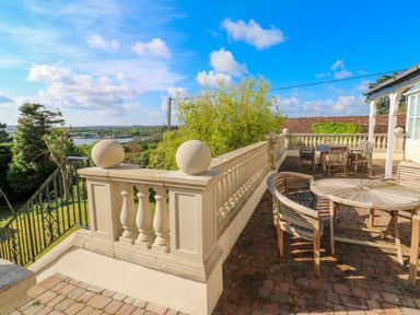 A patio with wooden tables and chairs and a stone balustrade overlooking a garden and river at Hindon Hall in Lelant near Carbis Bay