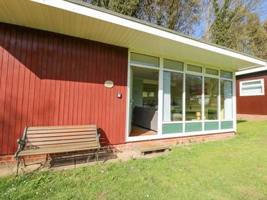 The exterior of a red wooden building with a bench and an open door showing a living area at The Beach Hut in Cromer