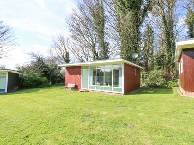 A small red wooden cabin with large windows and a bench outside on a grassy lawn with trees in the background at The Beach Hut in Cromer