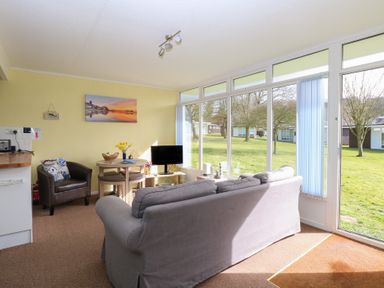 A living room with a gray sofa a round wooden table a leather chair and large windows overlooking a grassy area at The Beach Hut in Cromer