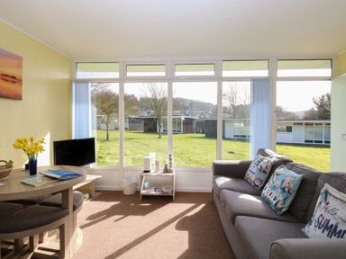 A living room with a gray sofa with decorative pillows a small wooden table with flowers a TV and large windows showing a lawn at The Beach Hut in Cromer