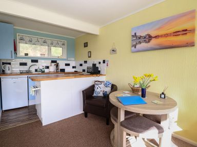 A kitchen area with blue cabinets a small fridge a round wooden table with chairs and a leather chair with cushions at The Beach Hut in Cromer