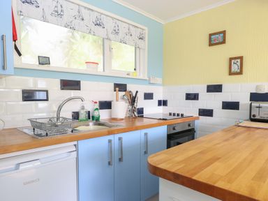A kitchen with blue cabinets wooden countertops a sink stove and a window with a sailboat patterned blind at The Beach Hut in Cromer