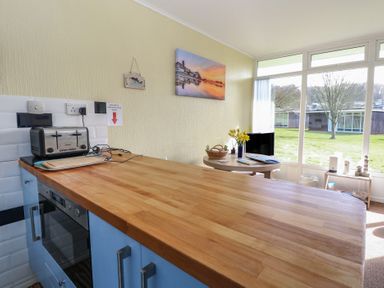 A kitchen counter with a toaster and cutting board overlooking a dining table with flowers and a television near large windows at The Beach Hut in Cromer