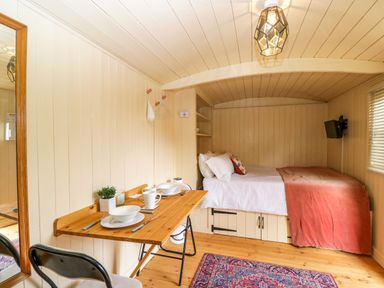 A small room with a wooden table set for two chairs a bed with white bedding and a red blanket under a curved ceiling at Mitford Gypsy Hut in Moreton-In-Marsh