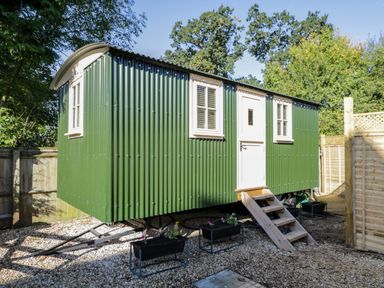 A green corrugated metal hut with white windows and door with wooden steps in a fenced garden area at Mitford Gypsy Hut in Moreton-In-Marsh