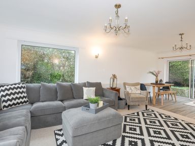 A living room with a grey sectional sofa and ottoman a beige armchair a dining table and chairs with sliding glass doors at 15 Maes Awel in Abersoch