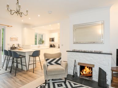 An open plan kitchen and living area with a countertop and bar stools a beige armchair by a fireplace with a fire burning at 15 Maes Awel in Abersoch