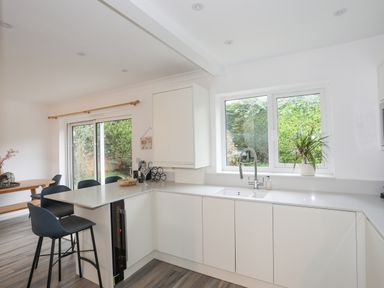 A kitchen with white cabinets a sink and a window with a plant on the sill and a dining area with black chairs at 15 Maes Awel in Abersoch