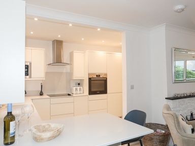 A modern kitchen with white cabinets appliances a countertop with a bottle of wine and wine glasses and an adjacent seating area at 15 Maes Awel in Abersoch