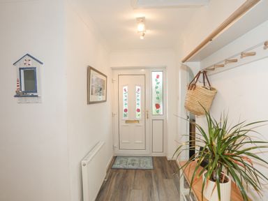 An entryway with a white front door with stained glass panels a wall-mounted coat rack with a basket a potted plant on a wooden bench a radiator a framed picture and a small mirror at 15 Maes Awel Abersoch