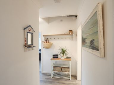 A hallway with a wooden shelf holding a woven bag and decorations a table with a plant and baskets and a framed painting on the wall at 15 Maes Awel in Abersoch
