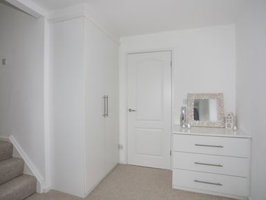 A white wardrobe and a white chest of drawers with a mirror on top next to a closed door at 15 Maes Awel in Abersoch