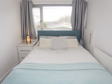 A bedroom with a bed with pillows and a blue throw blanket and a nightstand with a lamp at 15 Maes Awel in Abersoch