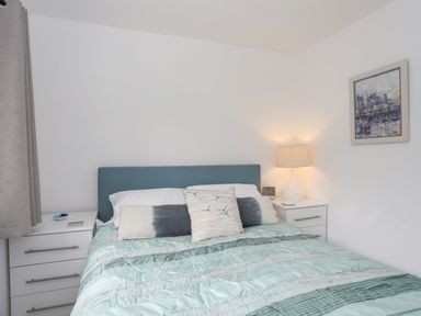 A bedroom with a bed with blue headboard and multiple pillows between two white bedside tables one with a lamp and a framed picture on the wall at 15 Maes Awel in Abersoch