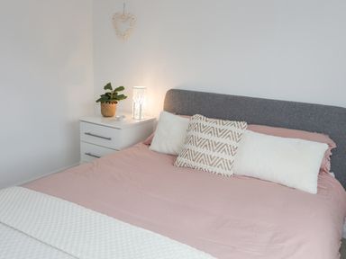 A bed with pink sheets two white pillows and a patterned cushion with a grey headboard a white bedside table with a plant and lamp at 15 Maes Awel in Abersoch