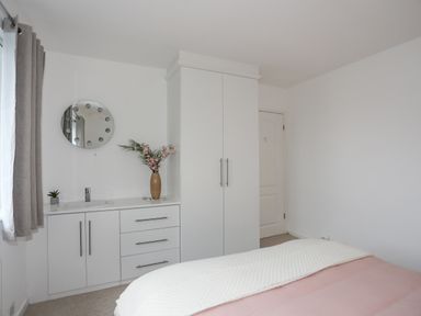 A bedroom corner with white built-in cabinets a round mirror flowers in a vase and a bed at 15 Maes Awel in Abersoch