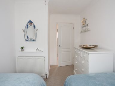 A bedroom with two beds covered in blue blankets a white dresser with a bowl of shells a boat-shaped mirror and a white door at 15 Maes Awel in Abersoch