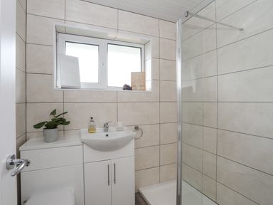 A bathroom with a white sink cabinet a potted plant a soap dispenser a shower with glass door and tiled walls at 15 Maes Awel in Abersoch