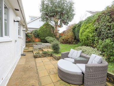 An outdoor patio area with rattan garden furniture and cushions next to a grassy garden with hedges at 15 Maes Awel in Abersoch