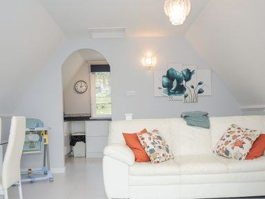 A living room with a white sofa with floral cushions and a folded blanket a clock on the wall and an archway leading to a kitchen at 24 Valley Lodge in Honicombe Park near Drakewalls