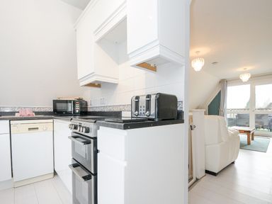 A kitchen with white cabinets a black toaster a microwave an oven and a dishwasher next to a living room with white chairs and glass doors at 24 Valley Lodge in Honicombe Park near Drakewalls