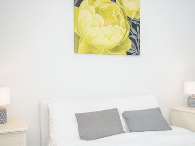 A bedroom with a bed white and grey pillows two bedside tables with lamps and yellow flower artwork above the headboard at 24 Valley Lodge in Honicombe Park near Drakewalls