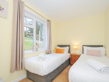 A bedroom with two single beds separated by a wooden nightstand with a lamp and a window with beige curtains at 24 Valley Lodge in Honicombe Park near Drakewalls