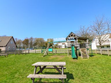 A playground with a wooden climbing frame and slide and a wooden picnic table on grass at 24 Valley Lodge in Honicombe Park near Drakewalls