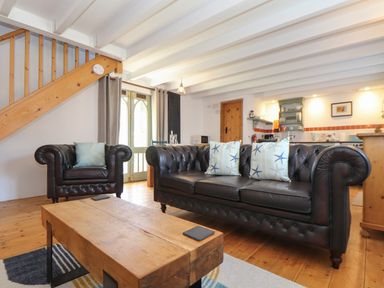 A living room with a leather sofa and armchair a wooden coffee table a staircase and a kitchen area at Oofoo's Barn in Mullion