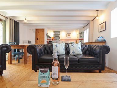 A living room with a black leather sofa and starfish cushions a wooden table with a bottle of wine a glass and a box of chocolates at Oofoo's Barn in Mullion