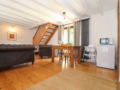 A living room with wooden floor leather sofas a wooden dining table with chairs a high chair a refrigerator a microwave a staircase and a large window with curtains at Oofoo's Barn in Mullion