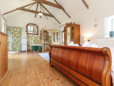 A bedroom with a wooden bed and ensuite bathroom with bathtub shower and sink at Oofoo's Barn in Mullion