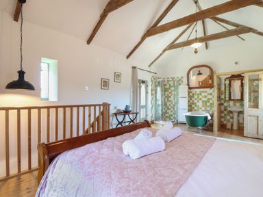 A bedroom with a wooden bed and rolled towels overlooking a bathroom with a freestanding bathtub and a tiled shower at Oofoo's Barn in Mullion