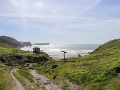 A coastal landscape with grassy hills dirt paths a wooden bridge a small hut a signpost and the ocean in the background at Oofoo's Barn in Mullion