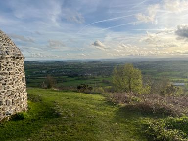 A grassy hilltop with a stone structure and a view of fields and trees at Piglet Corner in Culmstock