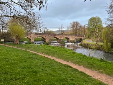 A stone bridge over a river with grass and trees on the riverbank at Piglet Corner in Culmstock
