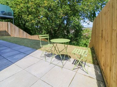 A patio with two green metal chairs and a green round table next to a wooden fence and trees at Valise in Bideford