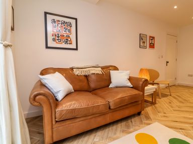 A brown leather sofa with two white pillows and a throw blanket in a room with framed artwork a wooden chair a side table with a lamp and wooden flooring at Valise in Bideford